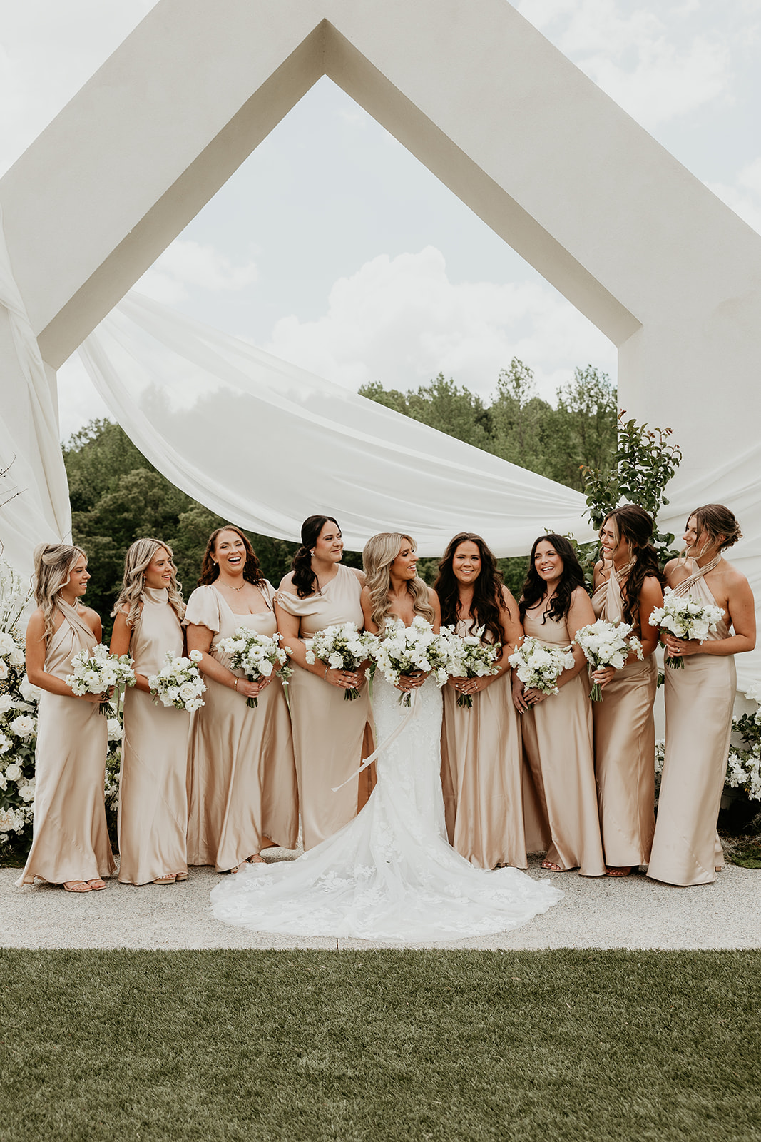 A bride and her bridesmaids posing in front of a modern wedding venue arch.