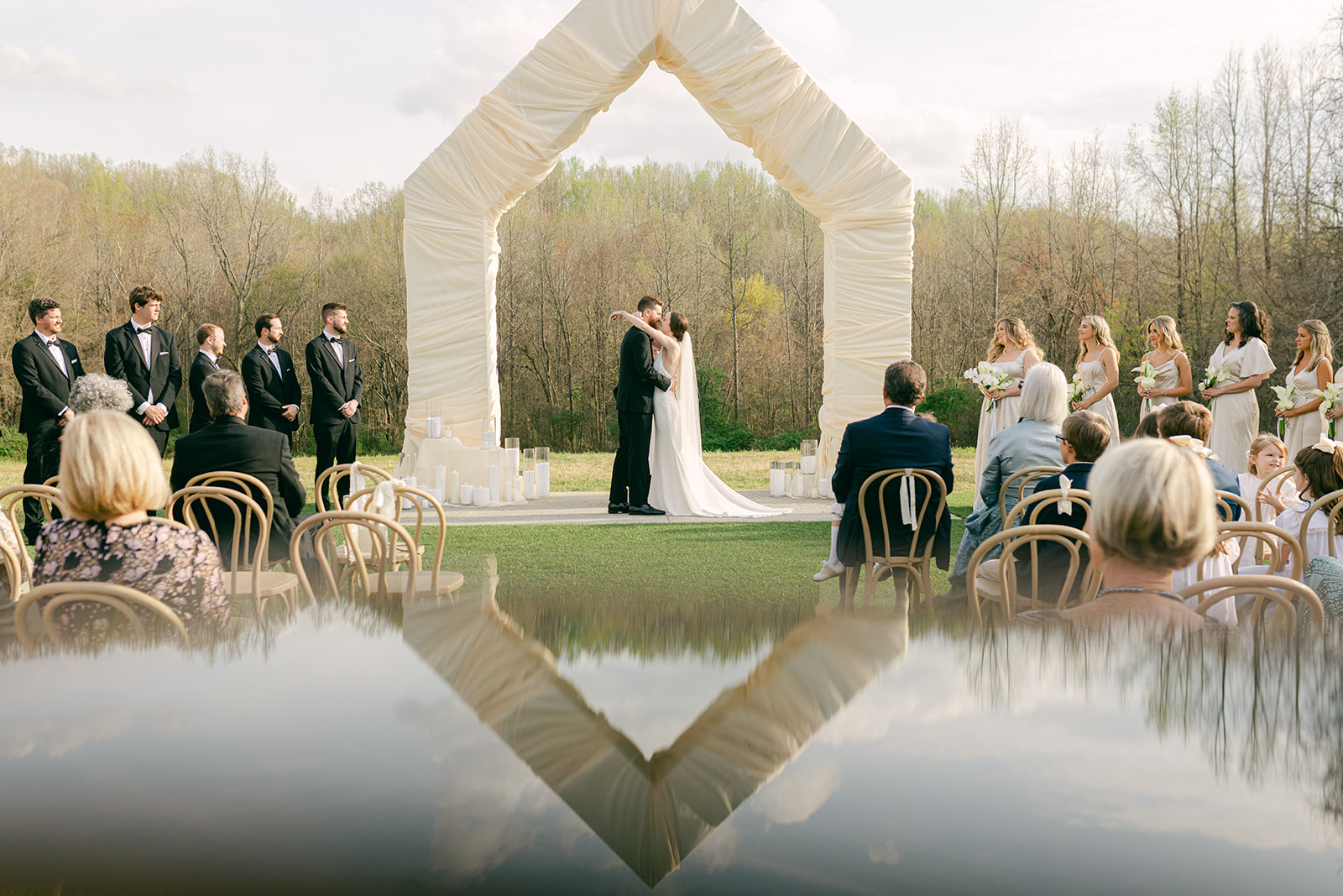 Bride and groom sharing their first kiss as husband and wife under a wedding arch in front of trees.