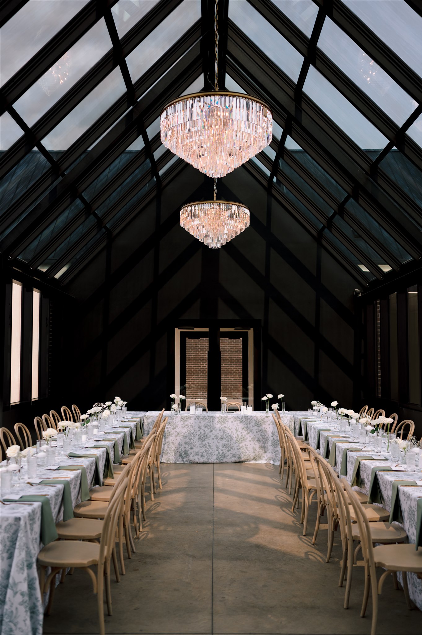 A modern glass house wedding venue with long tables and chandeliers.