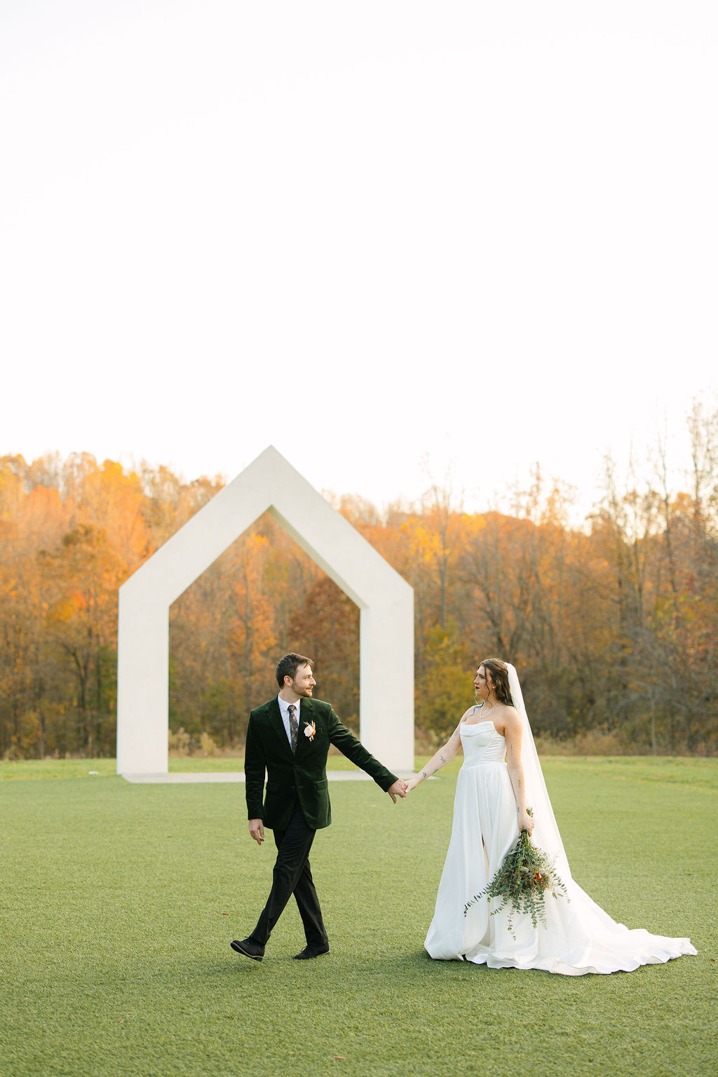 A bride and groom walking hand in hand in front of a modern wedding arch and fall colored North Carolina trees.