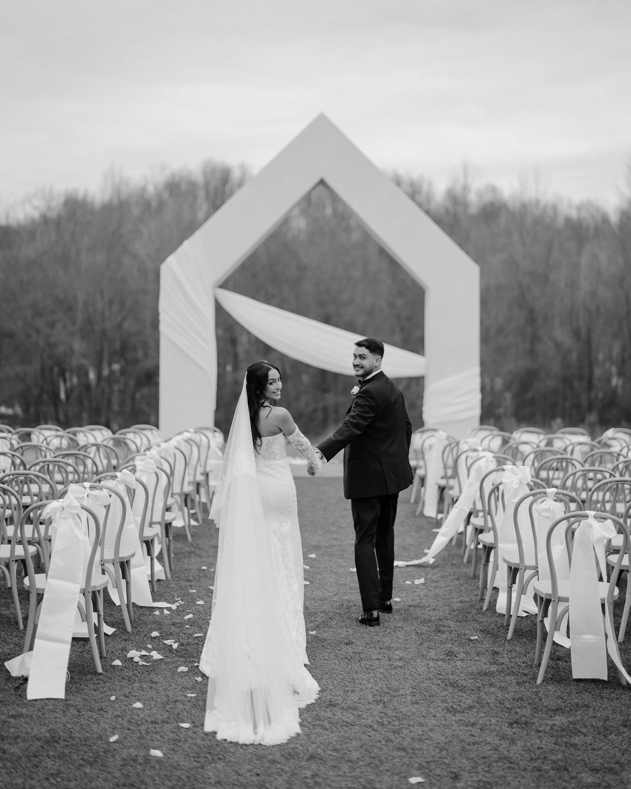 A bride and groom posing as they walk down their outdoor wedding aisle hand in hand.