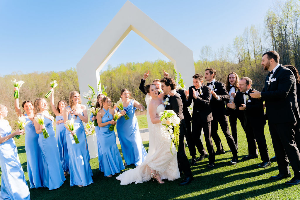 Bridal party posing in front of a modern wedding arch outside with blue dresses and black tuxes.