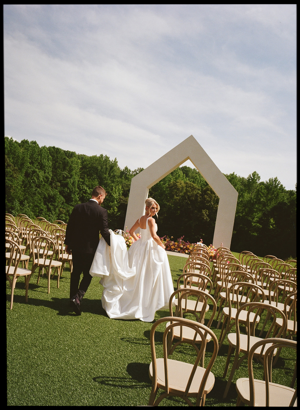 A groom carries his bride's wedding dress train as they walk through the aisle of their outdoor wedding ceremony.